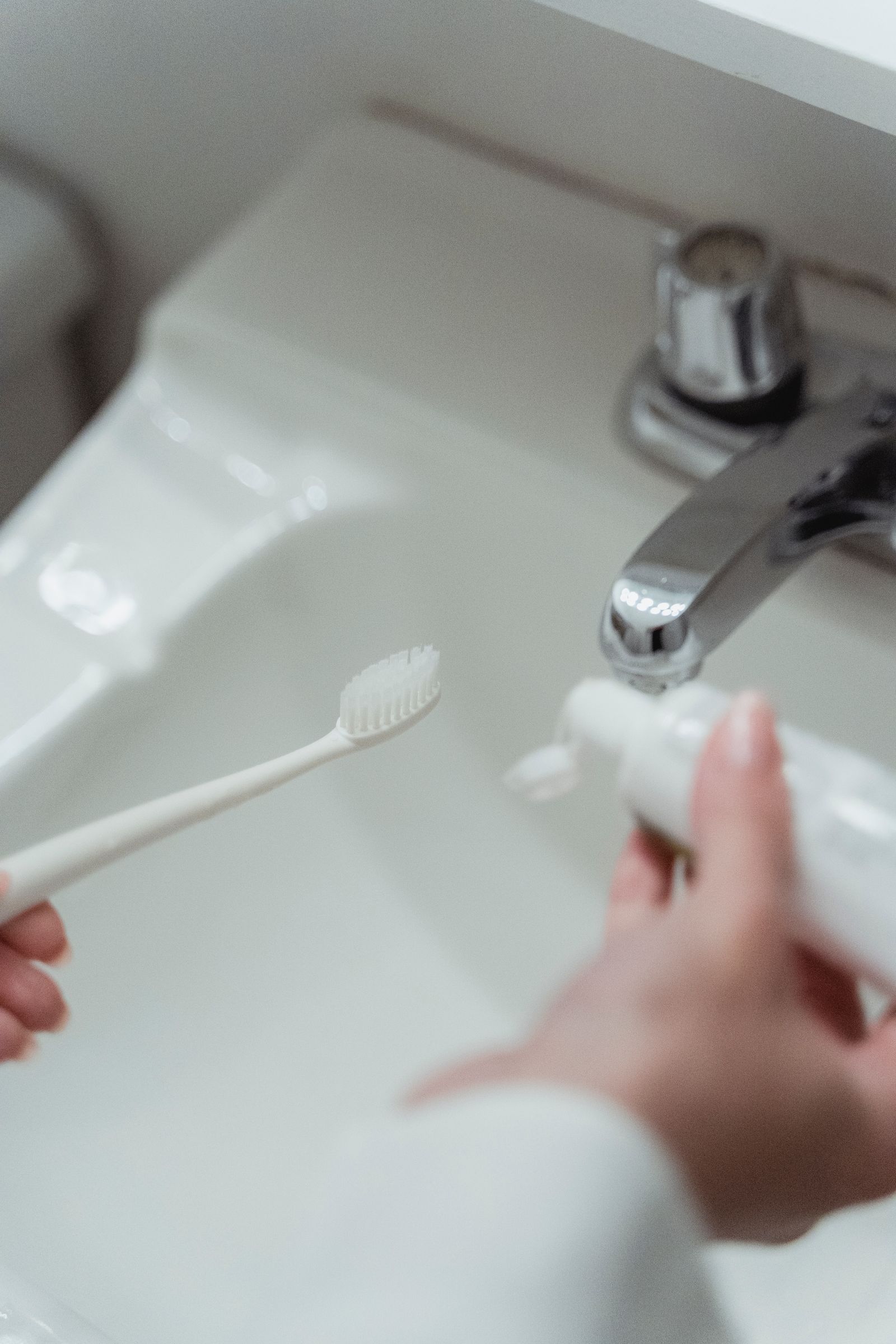 Overhead close-up of a hand holding a white toothbrush and toothpaste tube at a bathroom sink, no face visible